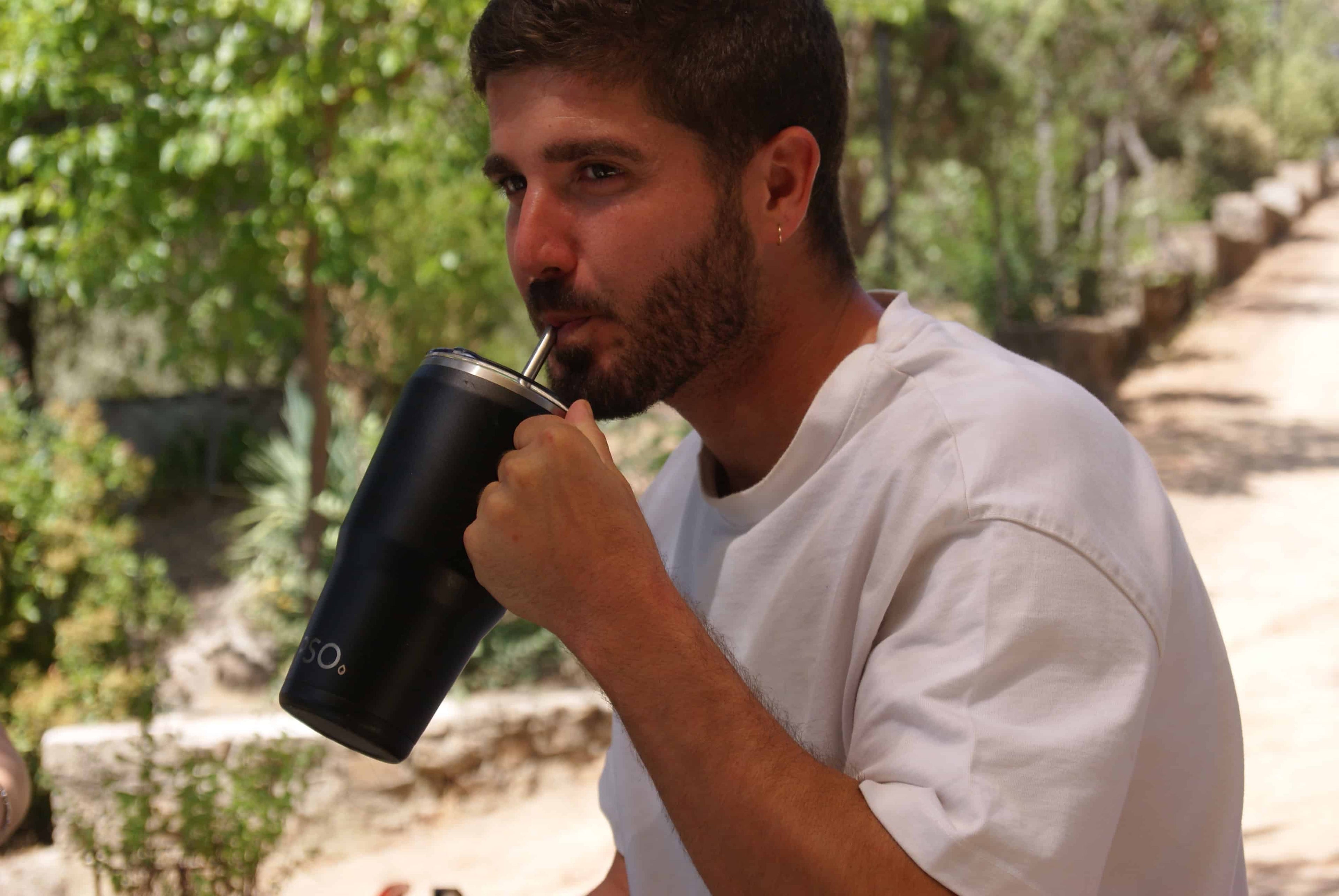 Hombre relajado en un chill out a la sombra, sosteniendo una taza térmica de acero inoxidable Dipso con un camino de tierra en el fondo.