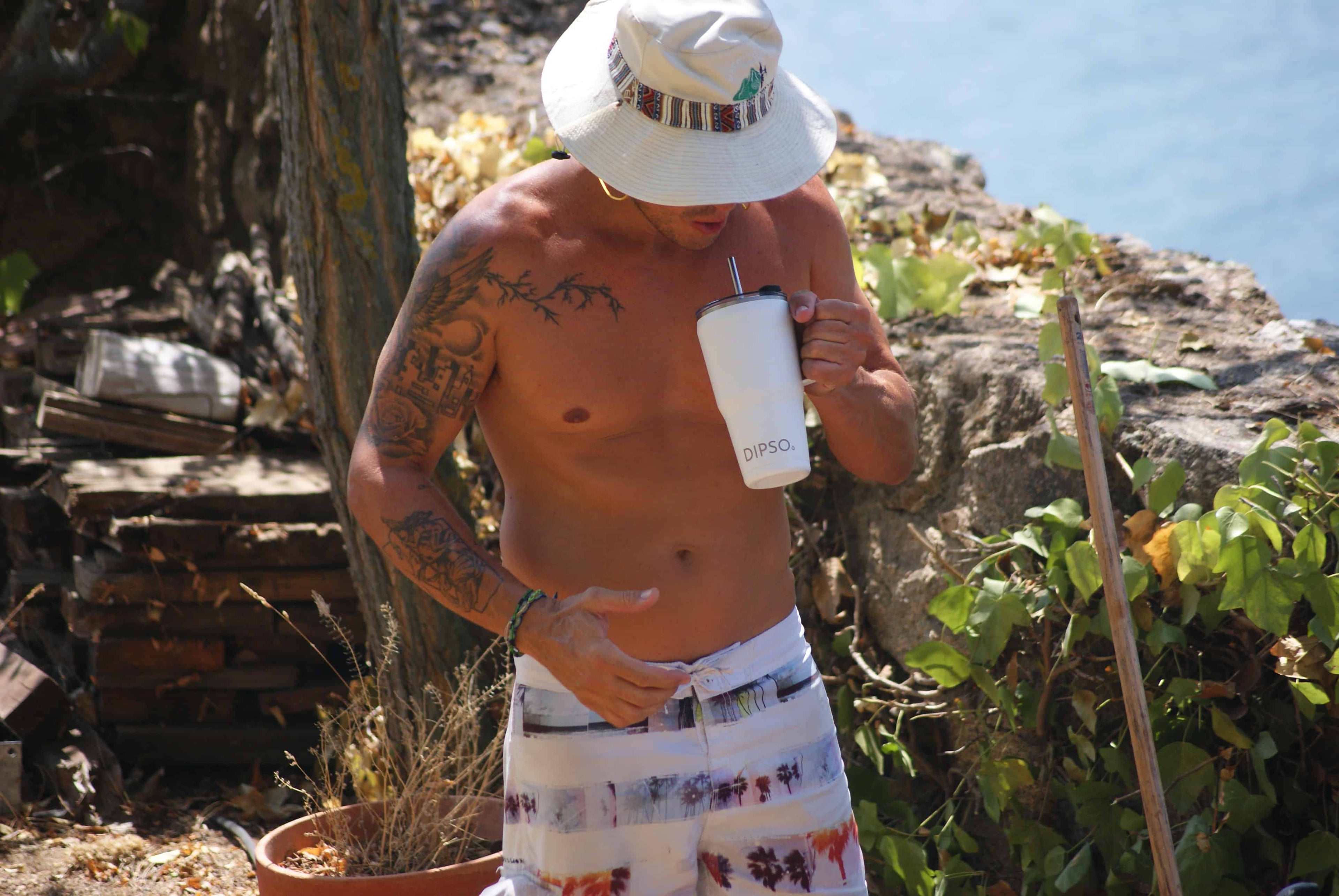 Hombre musculoso con tatuajes y gorro bebiendo de una taza térmica de acero inoxidable Dipso cerca del agua en un lago durante un día de verano.